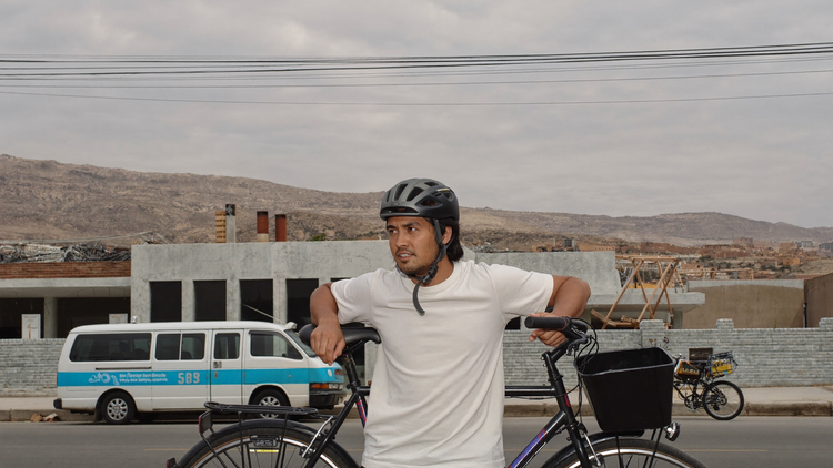 Ciclista peruano con casco puesto y correa abrochada junto a su bicicleta, calle de Lima con cerros al fondo